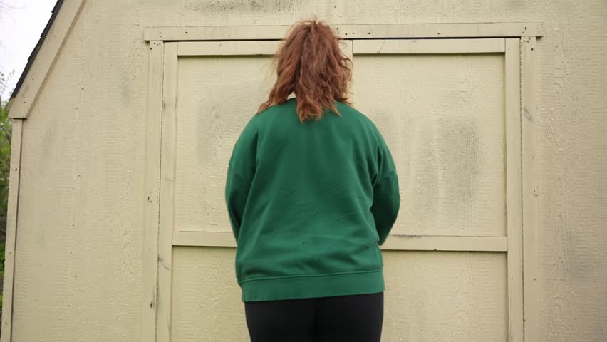 Woman entering a small outdoor storage shed or utility building, wearing a green sweater and headband in a casual backyard setting.