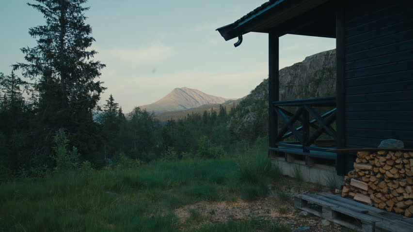 A traditional Norwegian mountain cabin overlooking a dramatic peak surrounded by forest and wilderness. Firewood stacked beside the cabin highlights authentic Scandinavian cabin life.