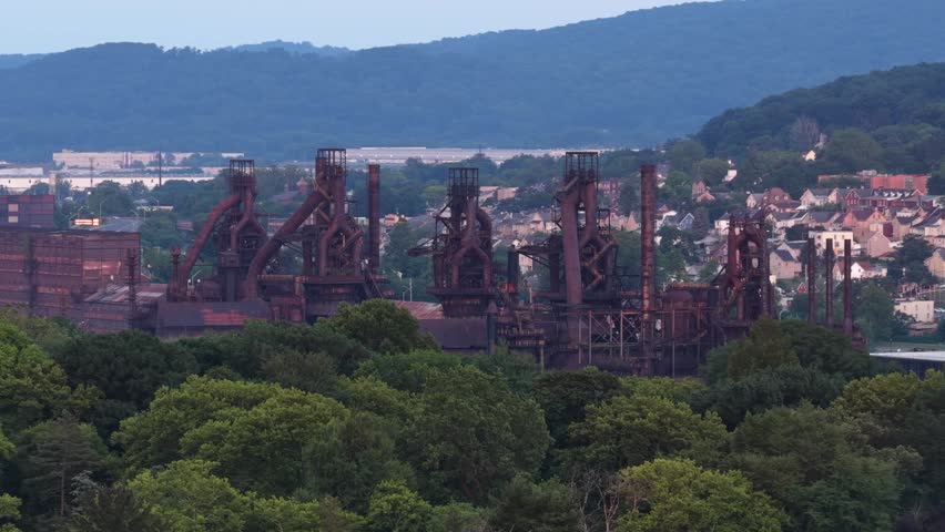 Row of rusted steel blast furnaces rises above dense tree line, historic industrial towers stand before residential rooftops and wooded hills in Bethlehem, Pennsylvania. Aerial drone wide shot