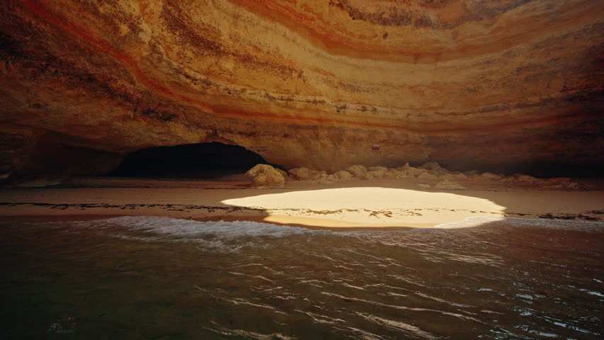 Sunlight illuminates a secluded sandy beach and layered limestone walls inside the famous Benagil Cave in Algarve, Portugal.