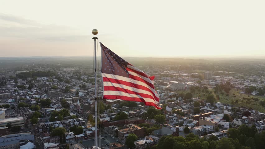 Giant American flag waves atop tall white flagpole above downtown Allentown rooftops, dense rowhouses and church steeple spread across hazy sunset skyline in Pennsylvania. Aerial drone close up