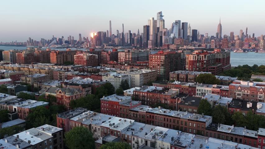 Brick brownstone rooftops fill Hoboken neighborhood, Manhattan skyline rises beyond Hudson River with Empire State Building glowing under golden sunset in New Jersey. Aerial drone flyover wide shot