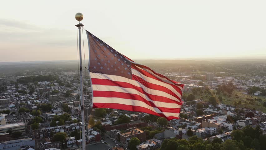 American flag waves atop tall flagpole above dense rowhome rooftops in small American town, warm golden sunset light spreads across quiet residential streets and trees. Aerial drone view.