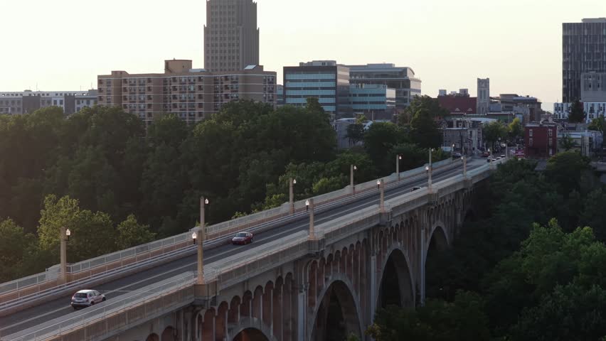 Cars cross historic Albertus Meyers Bridge above dense green treetops in Allentown, Pennsylvania, PPL Tower rises above hazy downtown city skyline at warm golden sunset. Aerial drone rising shot