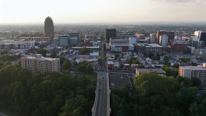 Elevated highway carries cars toward downtown Allentown, Pennsylvania, PPL Tower rises above dense office buildings and residential rooftops under golden sunset haze. Aerial drone approaching shot