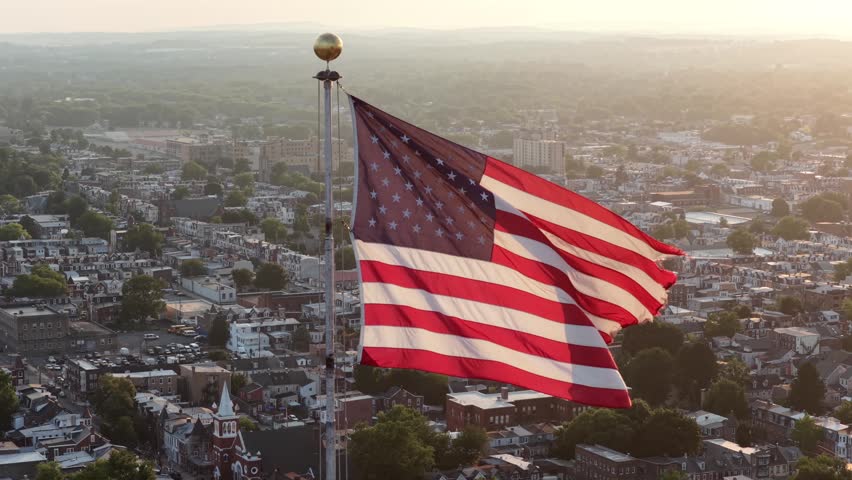 American flag waves above small town rooftops across United States, white church steeple and dense residential streets spread below warm golden evening sunlight glow. Aerial drone close up view.