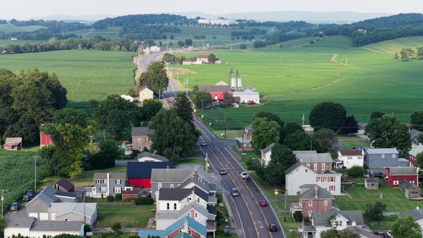 Red barn and grain silos rise beside curving country road through small farm town, cars drive past green cornfields and distant hazy rolling hillsides in rural America. Aerial drone wide shot