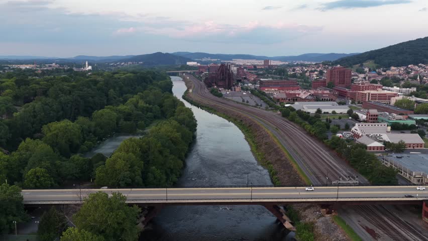 Highway bridge carries cars over Lehigh River near Bethlehem Steel Stacks in Bethlehem, Pennsylvania, railroad tracks and wooded hillsides spread beside quiet downtown. Aerial drone wide shot