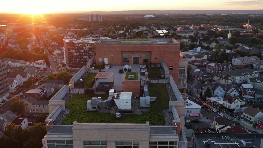 Luxury rooftop terrace with HVAC units and green plantings tops brick building in downtown New Brunswick, New Jersey, dense residential rooftops glow at golden sunset. Aerial drone flyover shot
