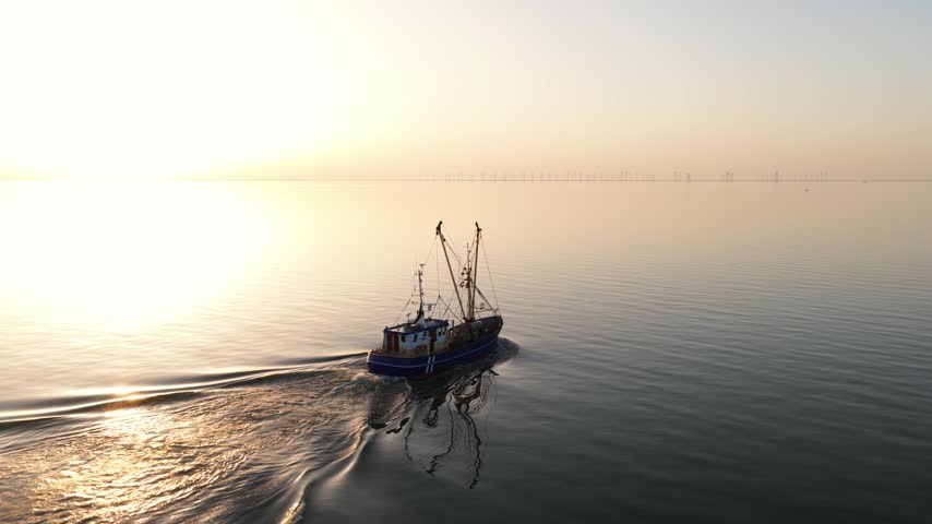 Aerial view of a fishing ship leaving the port of Stavoren, Friesland, during golden hour, with warm sunset light over the harbor and IJsselmeer waterfront.