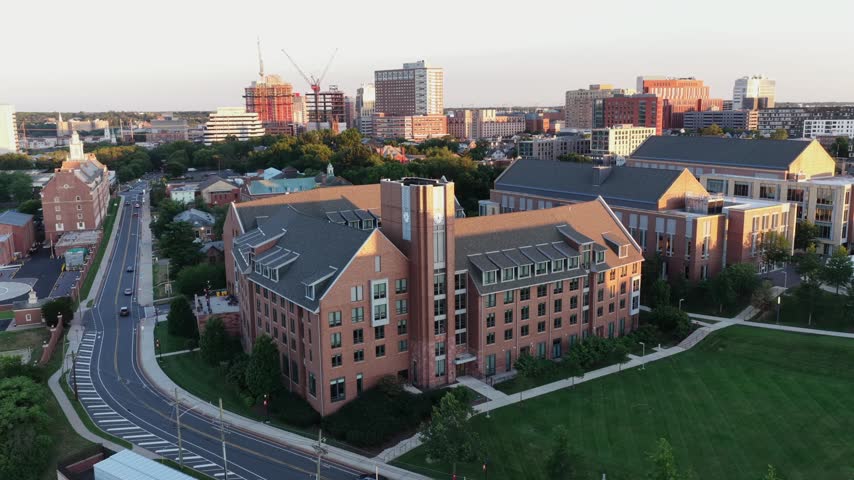 Car drives along curving road past brick dormitory building in New Brunswick, New Jersey, downtown skyline and construction cranes rise beyond green lawns at sunset light. Aerial drone view.
