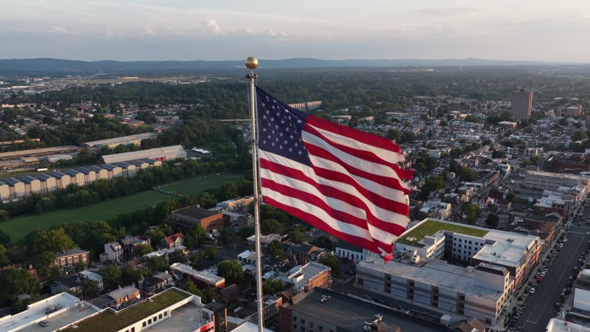Large American flag waves atop tall flagpole above downtown Allentown, Pennsylvania, dense rooftops and tree-lined residential streets spread across busy cityscape below. Aerial drone orbit shot