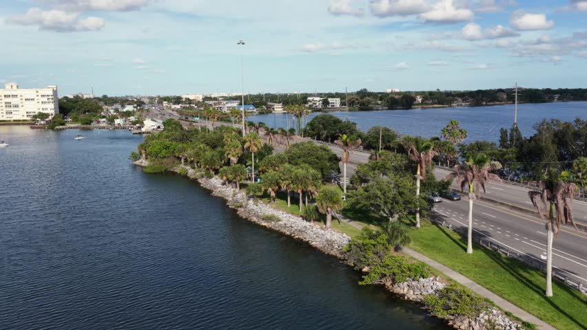 Palm-lined roadway curves along rocky waterway shoreline near Merritt Island Florida, cars travel busy highway beside calm blue water and distant apartment buildings. Aerial drone backwards shot