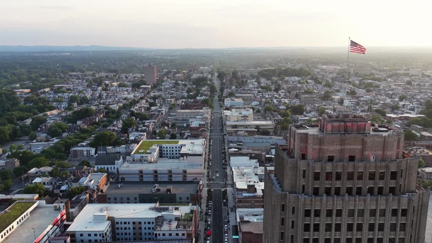 American flag flies above PPL Building in downtown Allentown, PA.Skyline at sunset, traffic moves along street through dense urban rooftops toward distant tree-lined hills. Aerial drone backwards wide