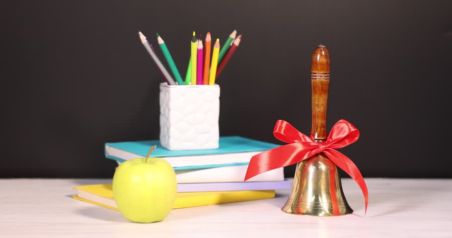 Woman putting flowers, school bell, books, apple and pencils at white wooden table near chalkboard, closeup