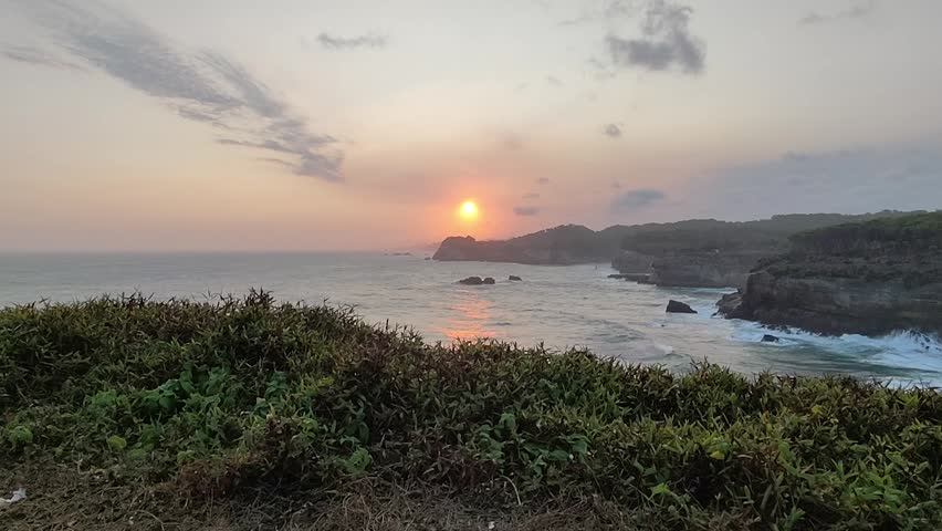 Sunset coastal landscape with rugged cliffs, ocean waves breaking against rocks, and green vegetation in the foreground under a warm orange sky with scattered clouds.