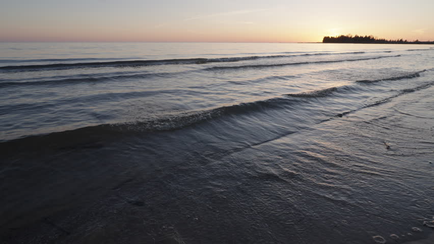Small waves lap gently onto the sandy shore of Lake Huron at sunset. The low-angle ground level perspective captures the orange sky reflecting on the wet sand on the Bruce Peninsula during spring.