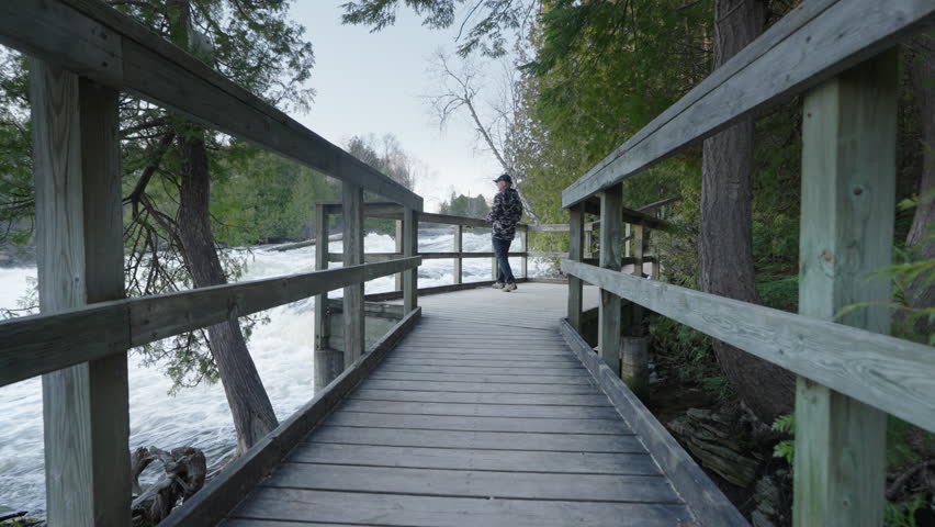 A wooden pedestrian bridge leads to a viewing platform overlooking the turbulent rapids of Sauble Falls. A person stands at the railing, watching the powerful spring river.