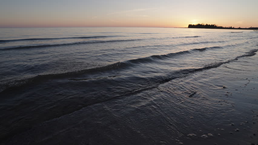 Soft waves gently wash over the flat sandy shoreline during a spring sunset on the Bruce Peninsula. The calm water of Lake Huron reflects the warm twilight sky on a quiet evening in Ontario.