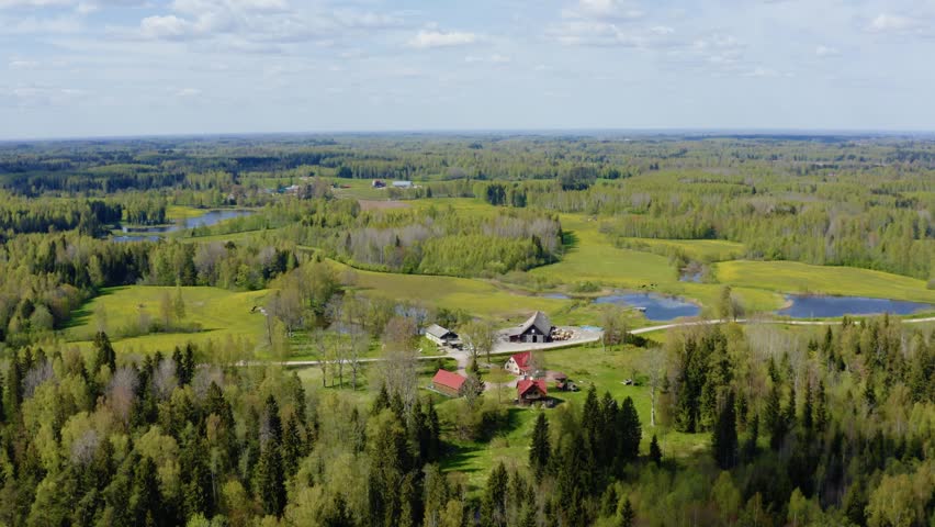 Aerial drone view of a rural farmhouse homestead nestled among lush green pine forests and lakes under blue sky.