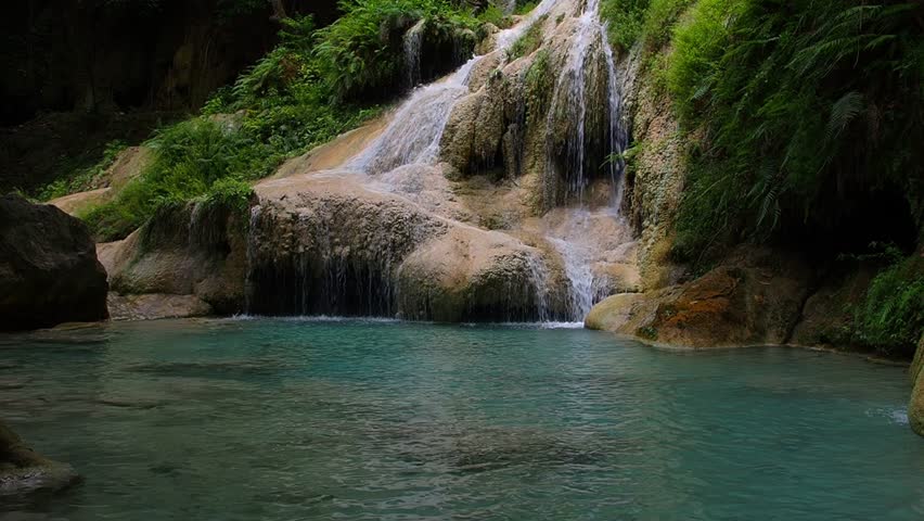 A waterfall with clear blue water, surrounded by natural rock formations and lush trees.