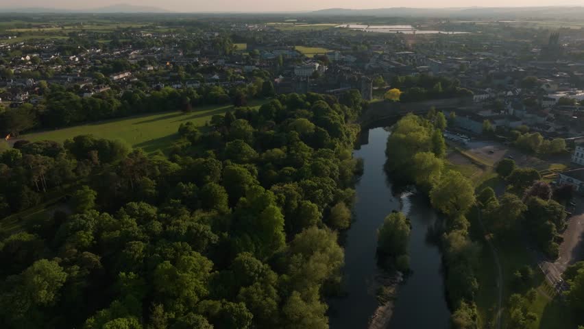 A cinematic aerial drone shot of Kilkenny city featuring the historic castle and River Nore during a warm sunset. Highlighting Irish heritage and urban nature.