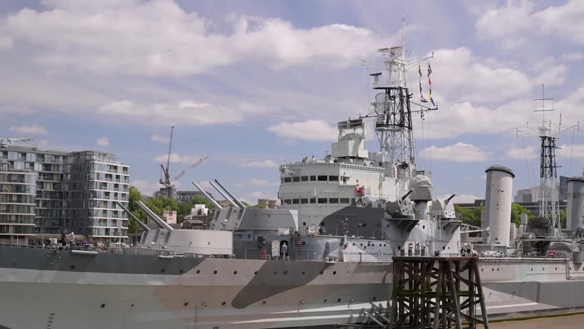 HMS Belfast World War Two cruiser sits alongside the quay with gun turrets and radar masts under a blue sky.