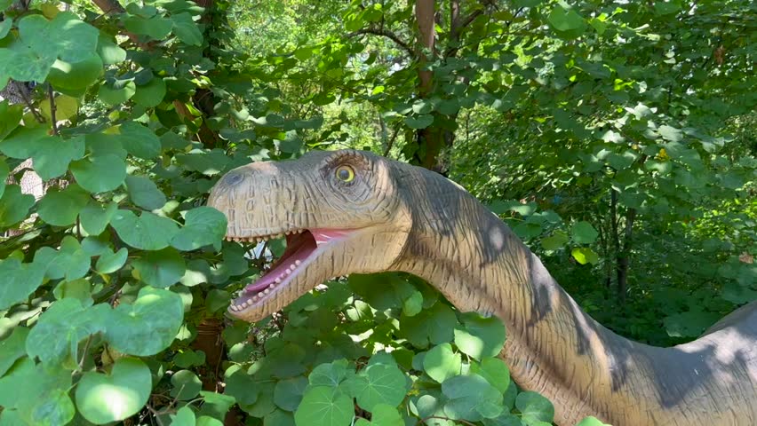 Long‑neck dinosaur statue with open mouth and teeth among dense leafy trees; dappled sunlight on the head and neck, tail visible at right. No text