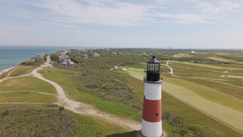 Sankaty Head Lighthouse red and white striped tower stands above coastal golf course fairways on Nantucket Island, sandy paths wind through dune grass beneath hazy sky. Aerial drone view.