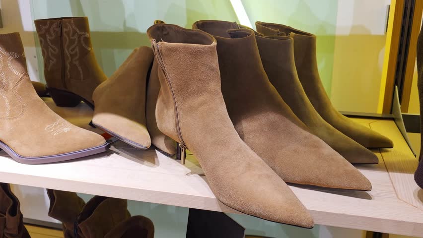 Close up view of multiple pairs of sophisticated tan suede ankle boots with zippers and low heels, arranged neatly on a light wood shelf within a contemporary shoe store, highlighting quality material