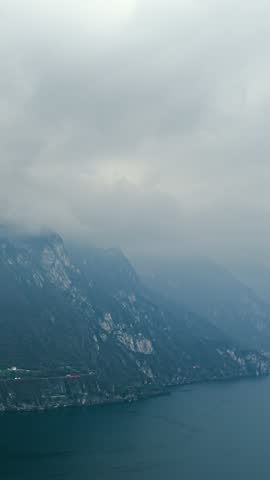 Timelapse shows low clouds drifting over steep misty cliffs and coastal road above calm lake water