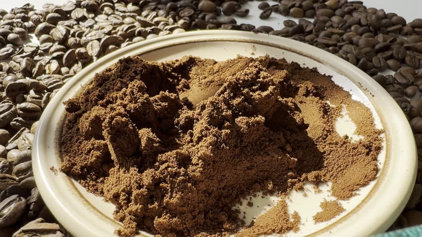 A plate among coffee beans. A spoon pours ground coffee onto the pile of ground coffee in the plate.