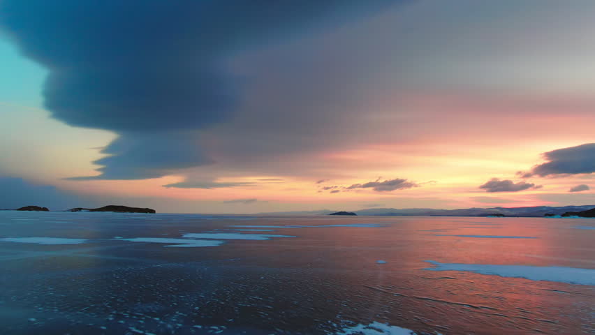 Beautiful sunset sky over frozen Lake Baikal. Icy landscape, snow cracks on winter ice water surface. Dramatic orange blue view reflection background. Nature scenery of Siberian wilderness.