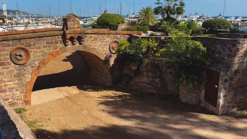 Historic stone arch bridge and fortified walls surrounded by Mediterranean greenery beside a yacht-filled marina in Antibes, France.