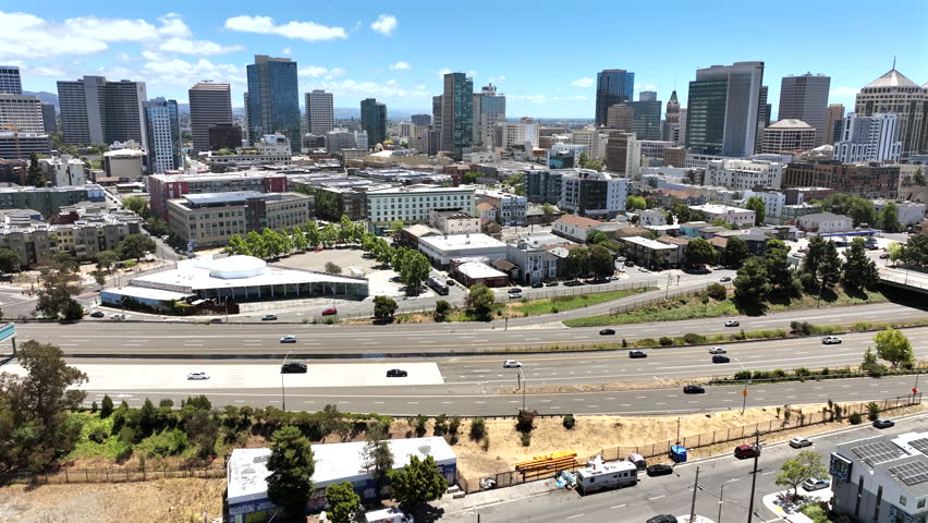 Wide aerial drone footage of Downtown Oakland on a bright sunny day, featuring the urban skyline, high-rise buildings, commercial properties, residential neighborhoods, busy streets, and vehicles