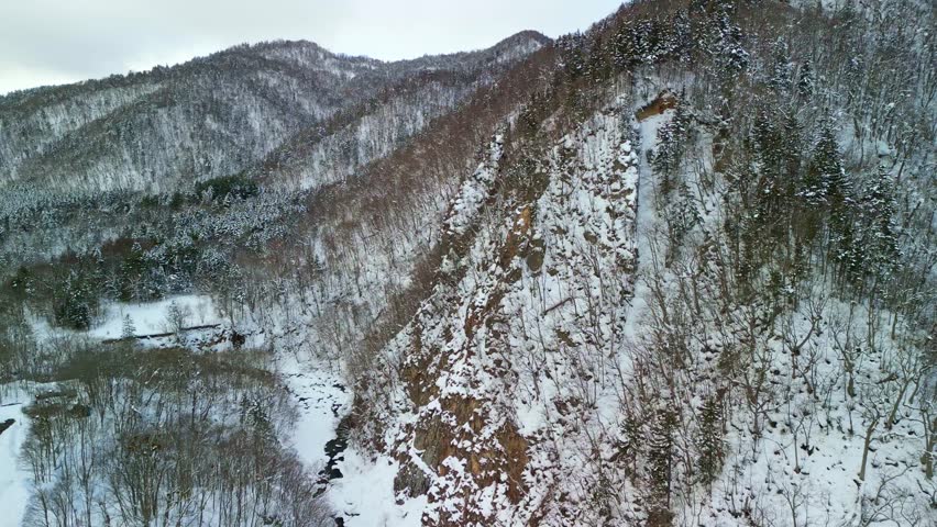 Aerial view panning across a dramatic snowy cliff side and river gorge leading to distant winter mountains.