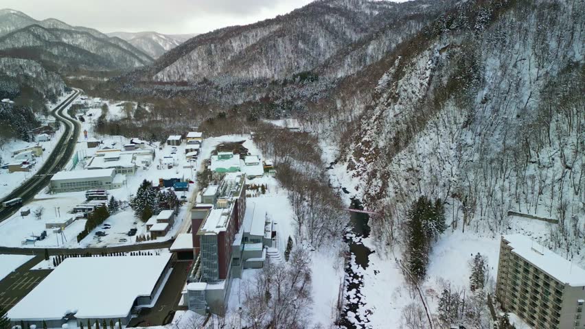 High drone view of a snowy mountain resort town with river and winter forest landscape in Hokkaido, Japan.
