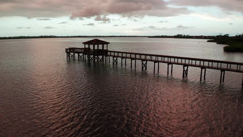 A covered pier stretches far into a wide inlet under pink twilight skies, with gentle ripples across the surface. Useful as a peaceful coastal establishing shot or background plate.