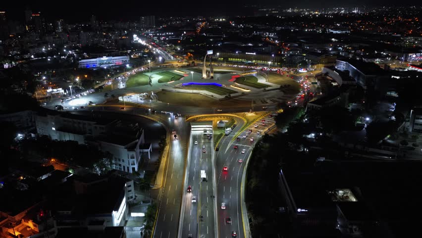Cars drive along curved highway toward Plaza de la Bandera monument in Santo Domingo, Dominican Republic, illuminated roundabout glows amid dense city lights at night. Aerial drone wide shot