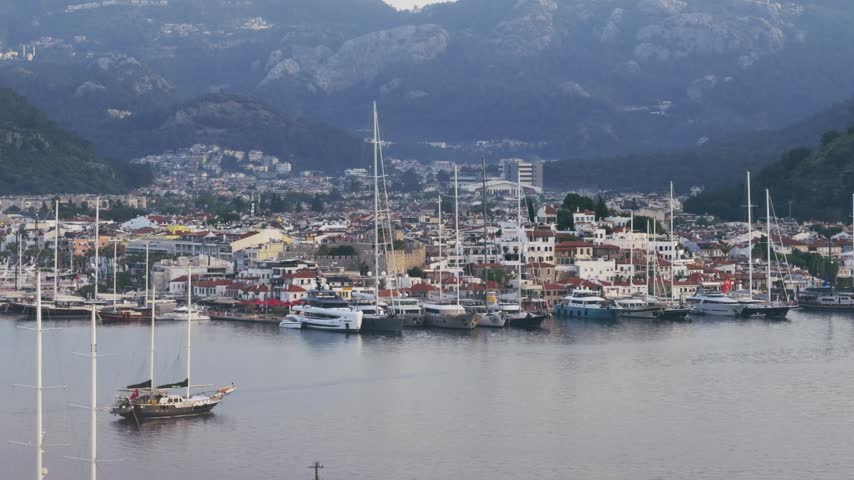 A serene view of boats and yachts moored at the historic Old Town pier in Marmaris, Turkey, against a backdrop of hills and buildings. Capturing authentic coastal life and travel.