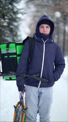 Young delivery courier in winter jacket with green backpack stands beside bicycle on snowy path in park, serious expression, cold weather.