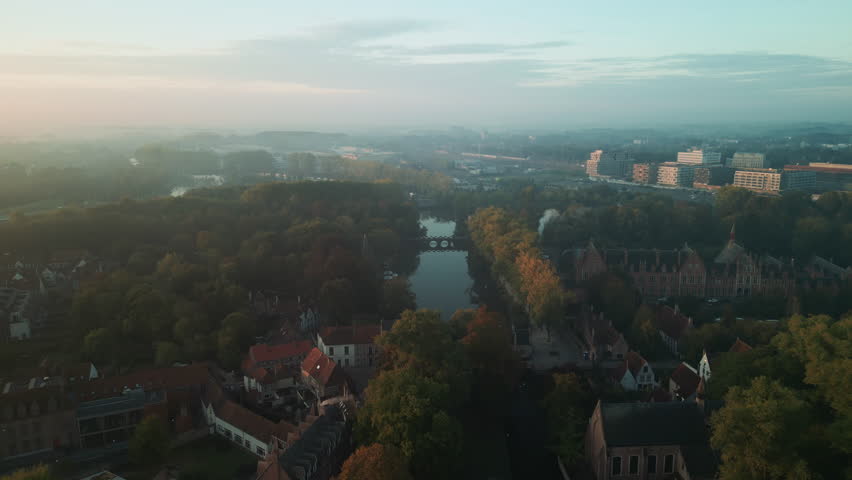Aerial drone view over a tree-lined canal in Bruges, Belgium, with autumn foliage, calm reflective water and the modern city district beyond morning haze.