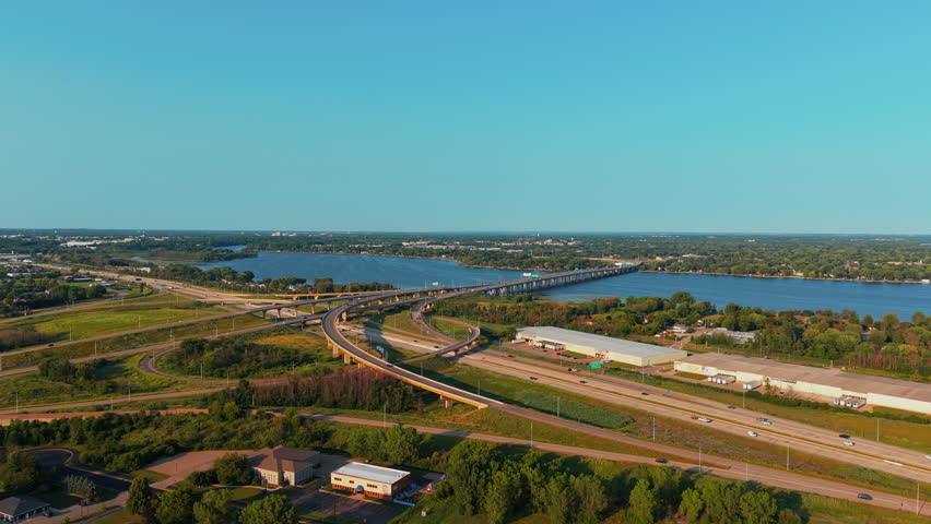 Aerial pull-back shot of sunlit highway 41 ramps curving toward a long bridge over Fox River beside suburbs and warehouses in Neenah, Wisconsin