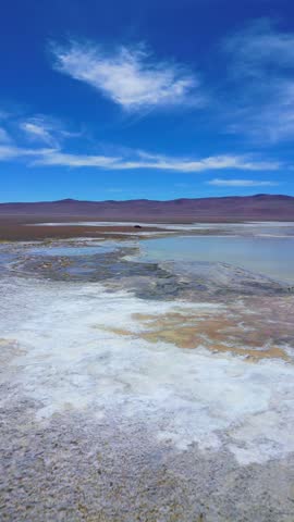 Forward flight over colorful mineral rich salt flat lagoon with textured shallow waters beneath desert mountains. San Pedro de Atacama, Chile