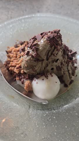 Japanese matcha shaved ice kakigori dessert with green tea ice cream topped with dark chocolate curls crunchy biscuit crumble and soft white mochi dango pearls in a textured glass bowl close up 360 view