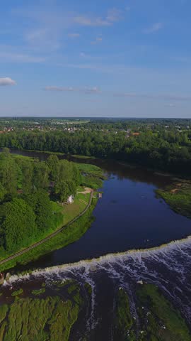 Aerial sequence shows Venta Rapid on the Venta River in Kuldiga, Latvia. White water spills over a curved ledge toward a dark pool, with paths, islets, and houses.