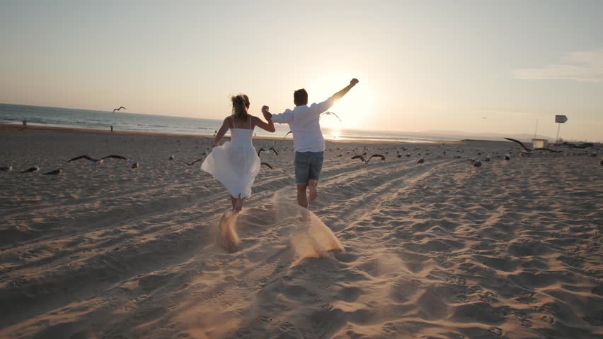 Couple runs on the beach at sunset while birds fly around them