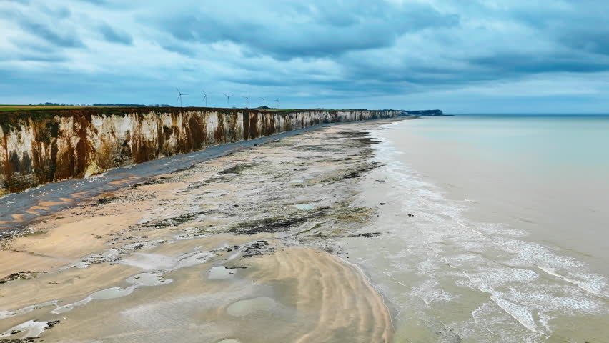 Normandy chalk cliff coastline above wide low tide beach on English Channel, France. Coastal travel landscape with wind turbines background