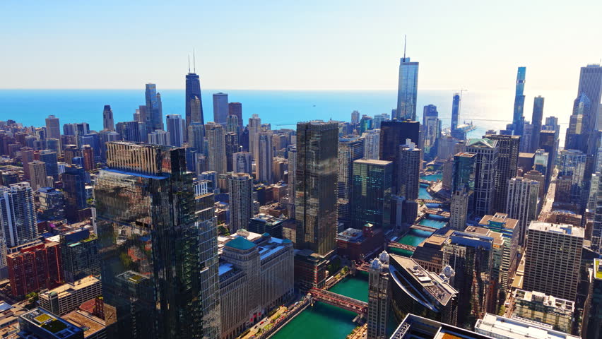 Sunlit skyline of Chicago and Lake Michigan, Illinois. Aerial panorama of downtown skyscrapers, river bridges and turquoise water beneath a clear blue sky.