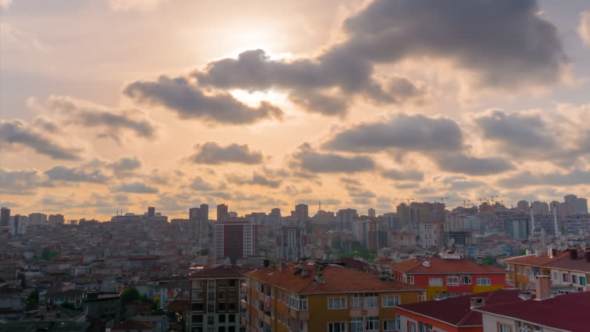 Timelapse: setting sun shining through fast moving clouds over Istanbul before dropping behind residential buildings. Orange sunset light is spreading over rooftops, getting darker - time lapse